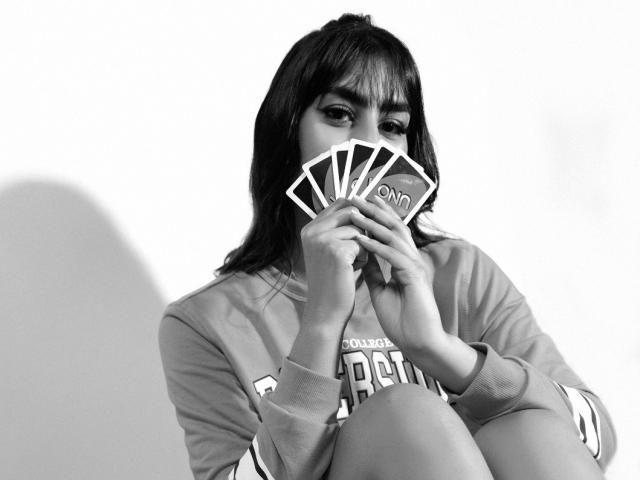 JessicaSoler, with dark hair and bangs, holding a fan of cards in front of her face, wearing a long-sleeve shirt, in a black-and-white photo.