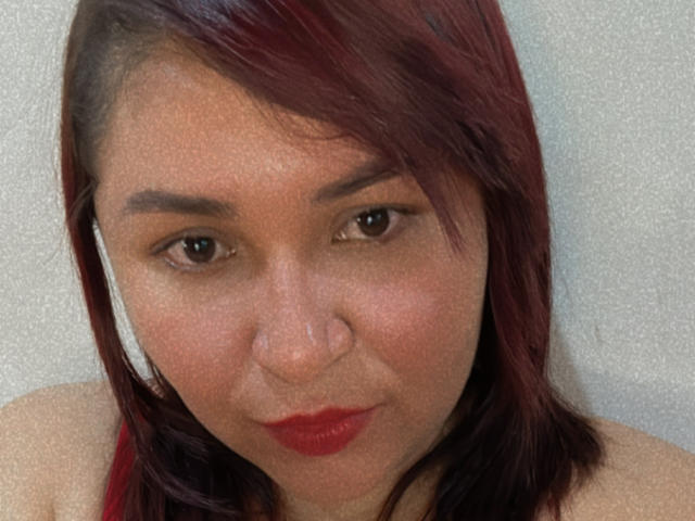 KarlaWild has medium brown skin, red lipstick, and dark red shoulder-length hair, looking straight at the camera with a slight, serious expression. She's wearing a red top.