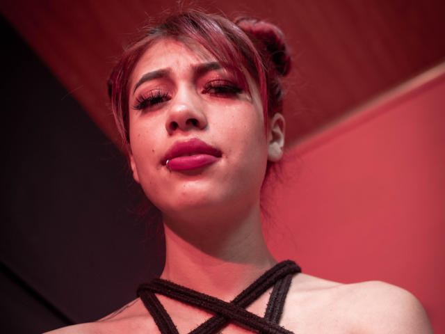 DalylaLove heeft roze lippenstift, rood haar in een knot, een zwart touw dat over haar borst loopt, en een licht geopende mond met een kleine zilveren piercing op haar onderlip..