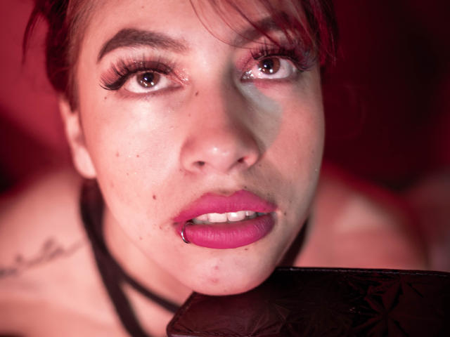 Le visage de DalylaLove, avec du rouge à lèvres rouge-rose, une lèvre inférieure percée, des sourcils foncés et de longs cils. Elle regarde vers le haut avec un léger rougissement, et porte un collier noir. Éclairage rouge..
