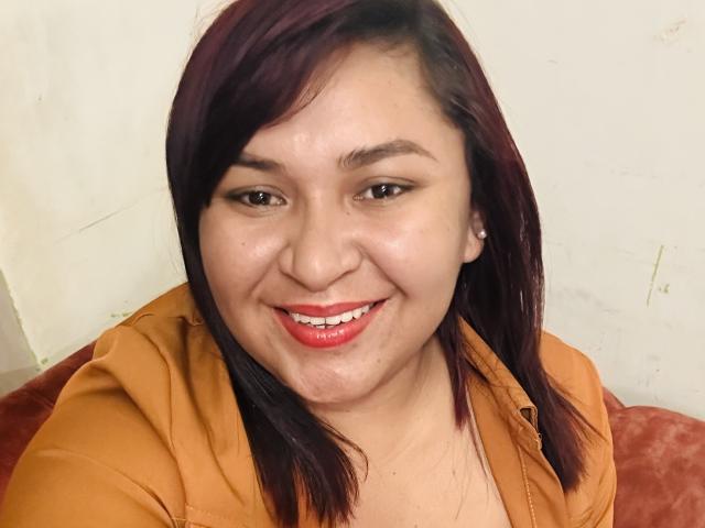 KarlaWild has dark red hair, light brown skin, and is smiling with red lipstick. She's wearing an orange hoodie, showing her tits slightly through the fabric.