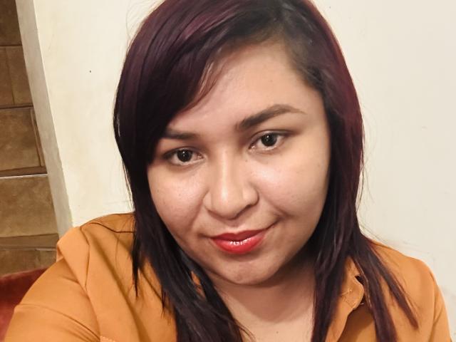 KarlaWild has dark brown eyes, light brown skin, and purple-dyed hair, wearing a shiny red lipstick and an orange shirt, smiling slightly at the camera.