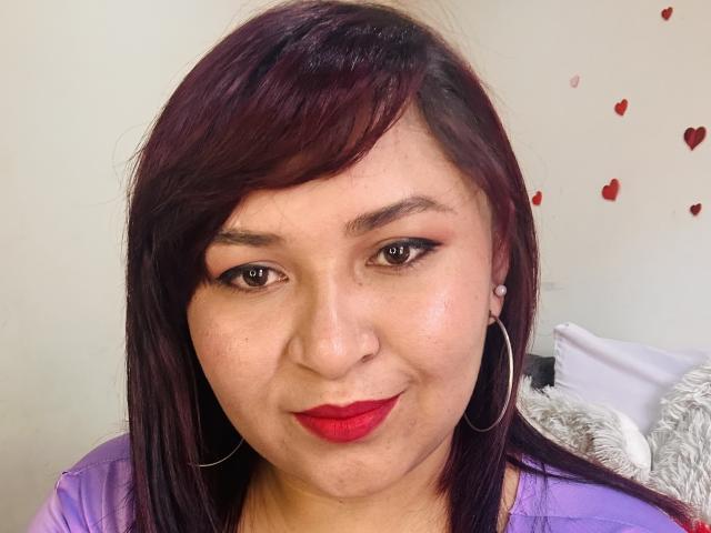 KarlaWild has dark purple hair, red lipstick, hoop earrings, and a nose ring. She's smiling with medium brown skin and subtle makeup. Her boobs are not visible.
