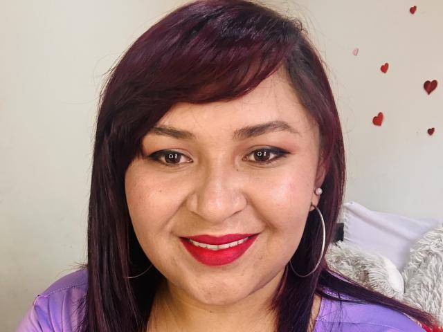 KarlaWild has red lipstick, dark purple hair, and hoop earrings, smiling with red-painted lips, wearing a purple top, showing her slightly chubby face and brown eyes.