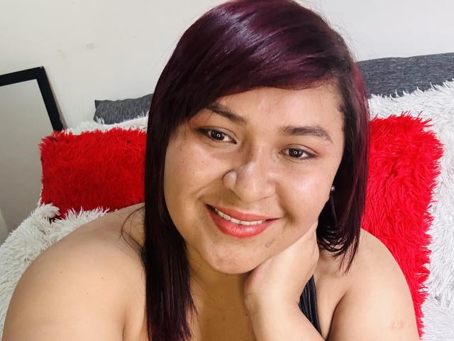 KarlaWild, com cabelo ruivo escuro, sorrindo, exibindo seus lábios rosados e seu nariz adorável, com a cabeça apoiada na mão, sem blusa, com uma alça preta visível..