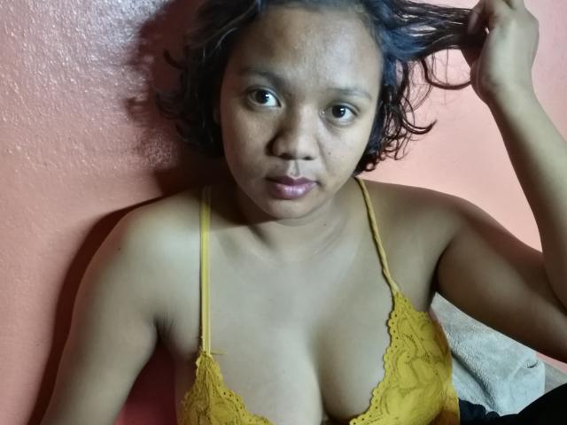 Morella69 with dark skin and short curly hair, wearing a yellow lace bra, holding her hair, looking at the camera with a neutral expression, small tits, pink wall.