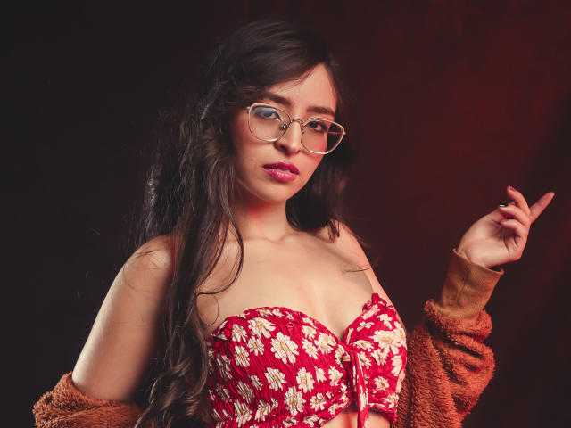 EvaMcAllister, avec de longs cheveux noirs, des lunettes, un bustier floral rouge, mettant en valeur ses seins, un cardigan marron duveteux, pointant avec sa main droite, fond rouge foncé, rouge à lèvres rose, expression confiante..