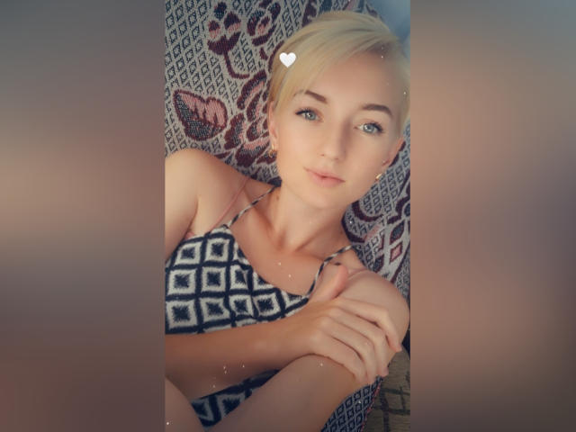 BeckiXJewel, con i capelli biondi corti, gli occhi azzurri e piccoli seni, indossa un bikini a pois neri e bianchi, tiene una mano al ginocchio, ha un'emoji a forma di cuore vicino alla testa e un sorriso dolce..
