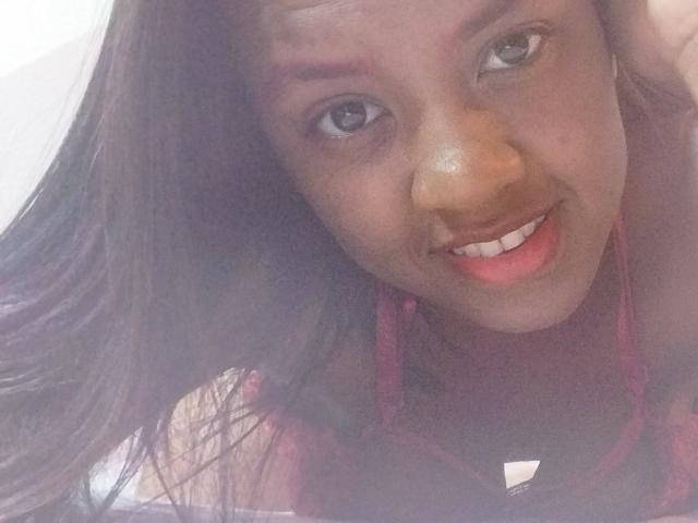 TiffanySammy with dark brown skin, long straight hair, smiling with pink lips, brown eyes, and red earrings, wearing a black top with a white triangle.
