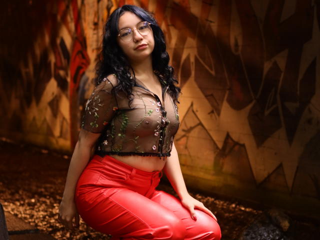 VeroHotLove con capelli neri ricci, occhiali, una maglia trasparente marrone con paillettes verdi, che mostra il seno, e pantaloni di pelle rossa, inginocchiata su un pavimento sporco, con un'aria sexy..