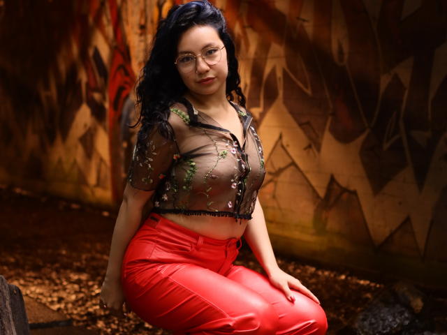 VeroHotLove with black curly hair, glasses, sheer black top with green floral embroidery, and red leather pants, kneeling on brown ground, showing her ass and medium tits.