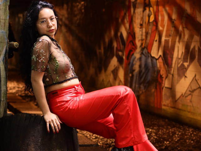 VeroHotLove with long black hair, glasses, sheer green-embellished top, red shiny pants, leaning on black cylinder, looking back with red lips, in dimly lit, rustic room.