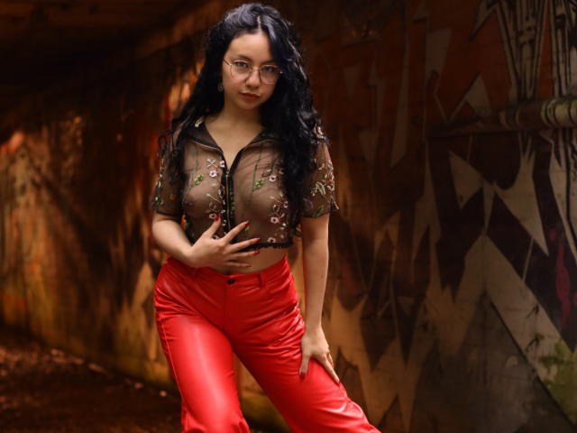VeroHotLove with black wavy hair, glasses, sheer floral top showing her boobs, red shiny pants, standing in a graffiti-covered, dimly-lit urban tunnel.