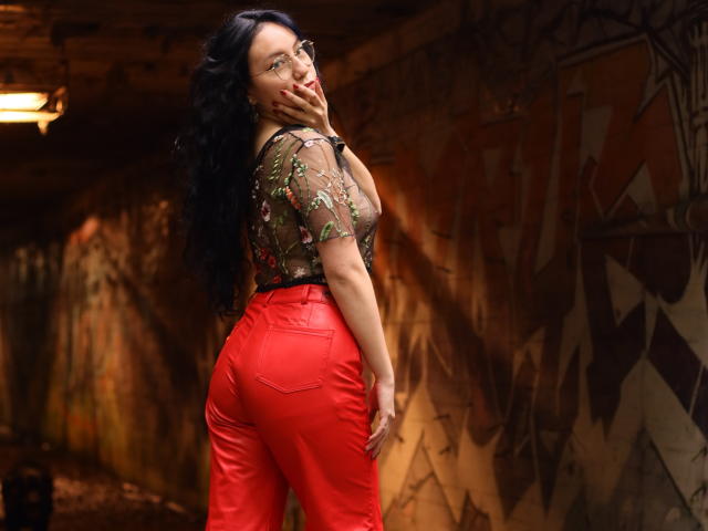 VeroHotLove with long black hair, glasses, sheer floral top, and bright red leather pants, showing off her ass, standing in a dimly lit, graffiti-covered room.