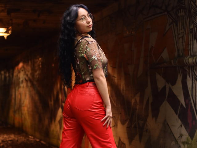 VeroHotLove, avec de longs cheveux noirs bouclés, portant un haut transparent à motifs floraux et un pantalon rouge moulant, debout avec ses fesses face à la caméra, regardant par-dessus son épaule..