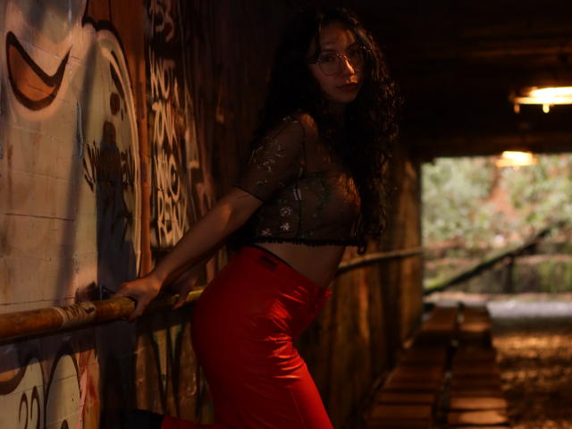 VeroHotLove with curly dark hair, wearing a sheer black top and red high-waisted pants, leaning on a railing in a dimly lit, graffiti-covered urban tunnel.