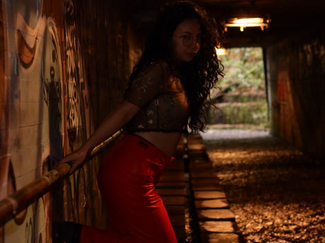 VeroHotLove tem cabelos pretos encaracolados, veste uma blusa preta transparente que deixa os seios à mostra, calças vermelhas de cintura alta e botas pretas, apoiada em um trilho enferrujado em um túnel com pouca luz..