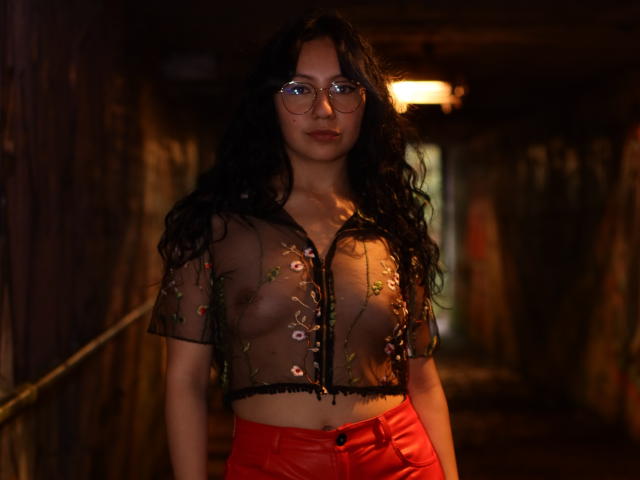 VeroHotLove, con cabello negro largo, gafas, una blusa transparente con bordados florales, pechos pequeños visibles y pantalones de cuero rojo, de pie en un pasillo de madera tenuemente iluminado..