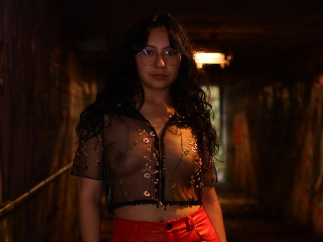 VeroHotLove mit langem, schwarzen, lockigen Haar, Brille und einem transparenten, schwarzen Oberteil mit Blumenmuster, das ihre kleinen, prallen Brüste zeigt, trägt rote, hochgeschnittene Hosen in einem schwach beleuchteten, hölzernen Flur..