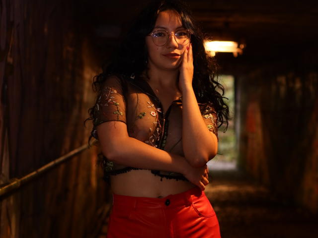 VeroHotLove con cabello negro y rizado largo, gafas, blusa floral transparente, mostrando pechos pequeños, pantalones rojos de cintura alta, tocándose la mejilla en un pasillo de madera tenuemente iluminado, con la mano en la cadera..