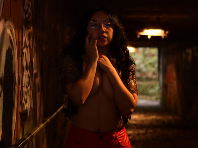 VeroHotLove, con cabello negro largo, gafas y pantalones rojos, sin top, cubriéndose los senos con las manos en un túnel tenuemente iluminado y cubierto de grafitis, mirando hacia abajo, con luz proveniente de atrás..