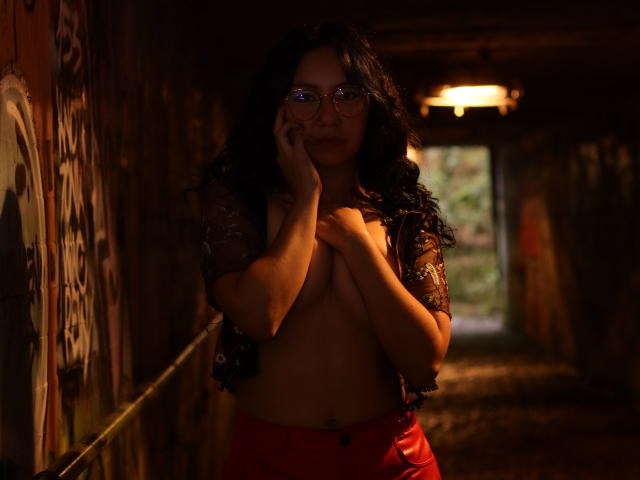 VeroHotLove mit dunklen, lockigen Haaren, die eine Brille trägt, ein durchsichtiges Spitzenoberteil und rote Hosen, steht in einem schlecht beleuchteten, mit Graffiti bedeckten Tunnel, bedeckt ihre Brüste mit den Händen und spricht am Telefon..