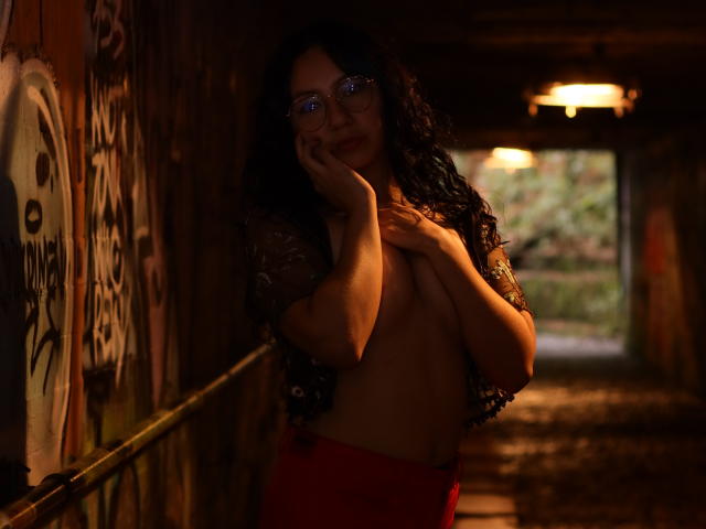 VeroHotLove, com cabelo preto encaracolado, óculos e saia vermelha, está em um túnel mal iluminado e coberto de grafites, cobrindo seus pequenos seios nus com as mãos, parecendo um pouco envergonhada..