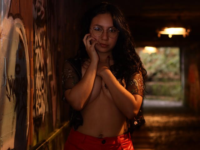 VeroHotLove, con i capelli neri ricci, gli occhiali e i pantaloni rossi, è in piedi in un tunnel poco illuminato, ricoperto di graffiti, e copre i suoi piccoli seni nudi con le mani, tenendo un telefono all'orecchio..