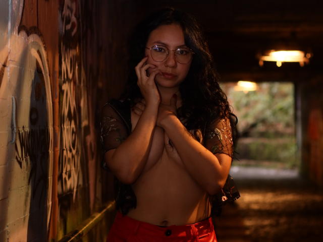 VeroHotLove, com longos cabelos cacheados pretos, óculos e calças vermelhas, está em um túnel coberto de grafites, cobrindo seus pequenos seios com as mãos, vestindo uma blusa preta transparente..