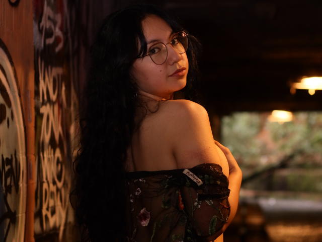 VeroHotLove with long black hair, glasses, and a sheer floral top, exposing her tits, leaning against a dark wooden wall, looking over her shoulder in a dimly lit room.