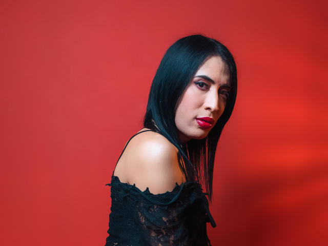 Antonella Humbolt, avec un haut noir en dentelle à épaules dénudées, du rouge à lèvres, et des cheveux noirs lisses, regardant par-dessus son épaule, sur un fond rouge, montrant son épaule nue et un maquillage subtil..