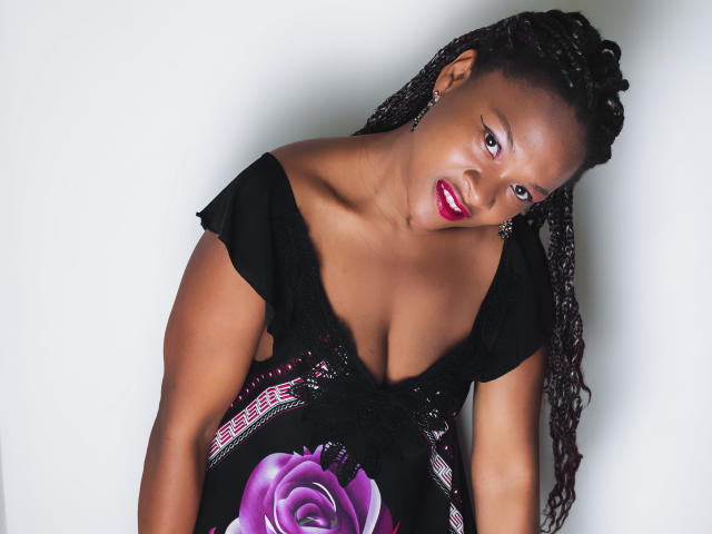 Camila Sandovali mit dunkler Haut, rotem Lippenstift und geflochtenen Haaren, trägt ein schwarzes Kleid mit einer violetten Rose, lehnt sich vor und zeigt ihre Brüste, während sie in die Kamera schaut..