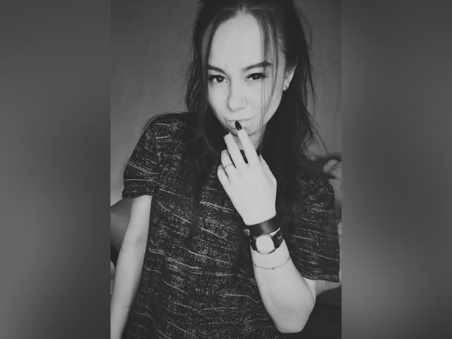 AlinaXRose con lo smalto nero sulle unghie, un dito sulle labbra, indossa una camicia scura con una texture particolare, un braccialetto al polso e lunghi capelli spettinati, e guarda seducentemente la fotocamera. Foto in bianco e nero..