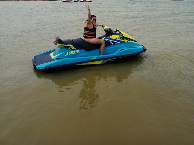 SharonKlart sur un jet ski bleu et jaune, portant un gilet de sauvetage noir et orange, avec son bras droit levé, assise sur l'eau et affichant un sourire joueur..