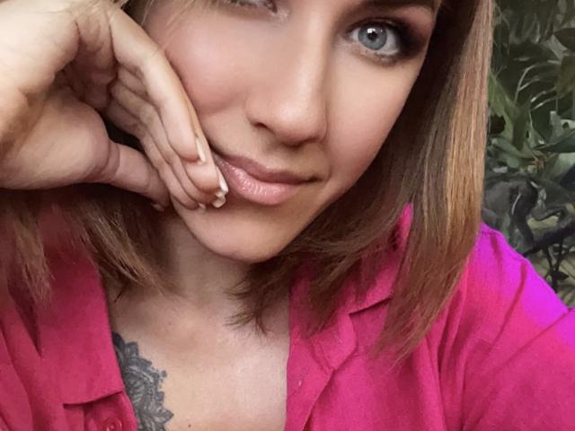 Misstyk with shoulder-length brown hair, blue eyes, and pink shirt, touching her cheek with her hand, showing a tattoo below her collarbone. Her lips are slightly glossy.