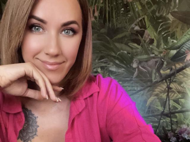 Misstyk tem um sorriso adorável, olhos verdes e um corte de cabelo bob castanho claro. Ela está vestindo uma camisa rosa, com uma tatuagem no peito, e apoiando o queixo na mão..