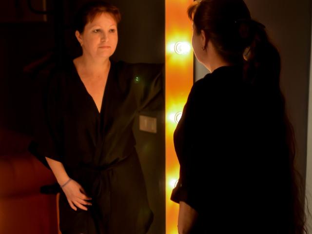 OVanessaHotly with red hair, wearing a black robe, stands in a dimly lit room, hands on her hips, her back to the camera, illuminated by warm light bulbs.
