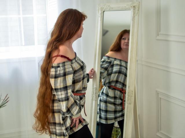 Vanessa Hotly, com cabelos ruivos longos, camisa xadrez sem mangas, sutiã vermelho visível, calças pretas, em pé em frente a um espelho, refletindo suas costas e nádegas..