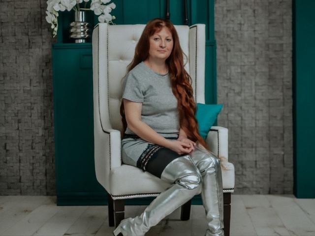 OVanessaHotly with long red hair, gray shirt, black lace skirt, and shiny silver thigh-high boots, sitting on a white upholstered chair, looking slightly bored.