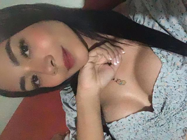 Katalina Lopez tem cabelos longos e escuros, pele morena e está vestindo um vestido floral com um decote baixo, que mostra seus seios. Ela está tocando o pescoço com um colar de ouro delicado..