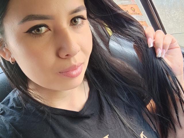 AshleyKatelyn has tan skin, long black hair, and is wearing a black shirt, with a slight pout, holding her hair, showing off her manicured nails with pink polish.