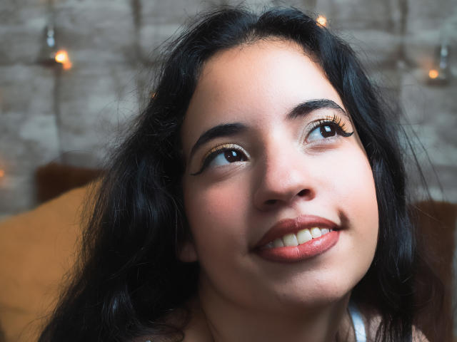 NicolSky tiene el cabello oscuro y ondulado, ojos marrones con delineador dorado, y una sonrisa encantadora con labios rosados. Está mirando hacia arriba con una expresión suave y feliz..