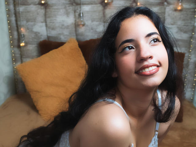 NicolSky with long black hair, smiling up, brown eyes, wearing a white lace top, showing her cute face and small tits, on a brown couch with yellow pillows.