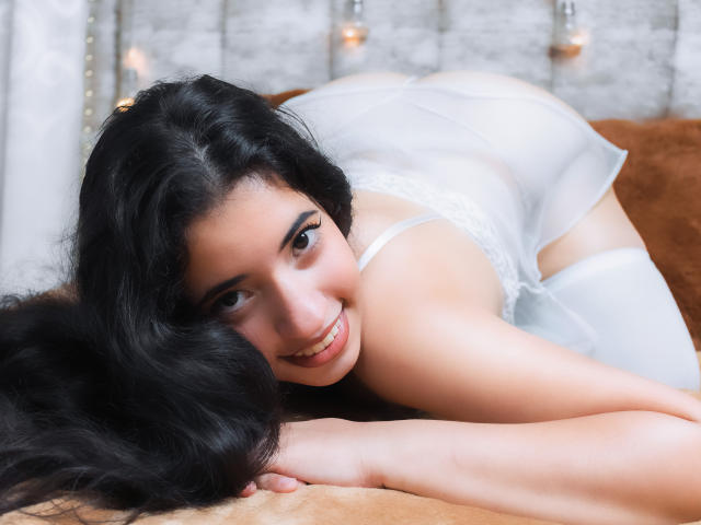 NicolSky with long black hair, smiling, wearing white lingerie, on her knees, butt up, hands on a brown surface, looking at the camera, soft light, warm brown background.