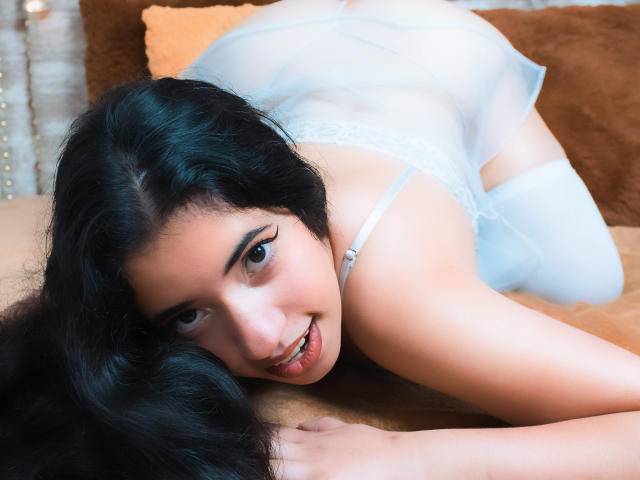 NicolSky, avec ses longs cheveux noirs, sa lingerie blanche et ses bas hauts, à genoux, regardant l'appareil photo avec un sourire, ses fesses relevées, les mains sur une surface marron..