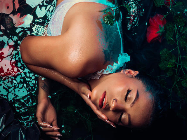 Amberv's face is glistening, eyes closed, with a hand touching her cheek. She's wearing a white, lace top, and has red lipstick, with green leaves and flowers around her.