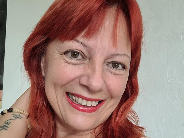 BarbaraBowie with red hair, smiling, showing her teeth, pink lipstick, tattoo on shoulder, white textured wall in background, wearing a bead bracelet.