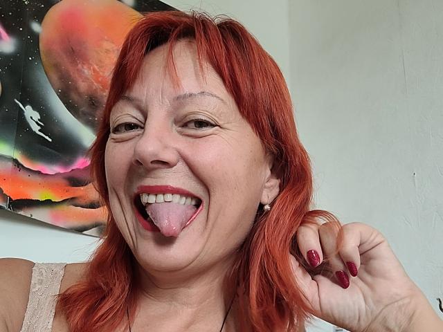 BarbaraBowie with red hair, smiling, tongue out, red lipstick, red nails, wearing lace top, holding hair, space painting in background.