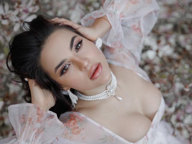 Celeste Belucci con un vestido transparente con estampado floral, las manos en el cabello negro, ojos brillantes, collar de perlas, escote, aretes blancos, acostada sobre pétalos rosas y blancos, mirando hacia arriba con los labios entreabiertos..