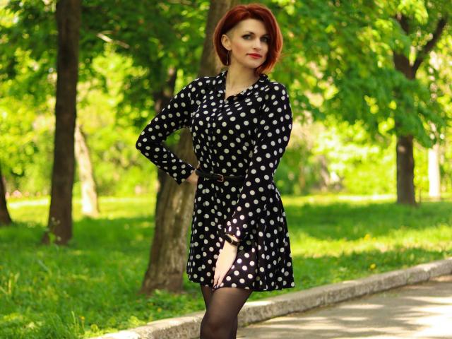 LisaTreds hat rote Haare und trägt ein schwarzes Kleid mit weißen Punkten und einem Gürtel, schwarze Strumpfhosen. Sie steht mit einer Hand in der Hüfte in einem grünen Park..